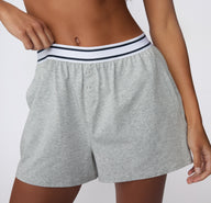 On a white studio backdrop, a model wears the CloudCotton Short in Heather Grey, showing the relaxed fit, soft cotton fabric, and elastic waistband with a button-front detail.