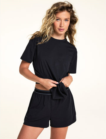 In a studio setting, Alexis wears the Cloud Cotton Shorts in Black with the matching Cloud Cotton Tee, featuring a relaxed fit, soft fabric, elastic waistband with contrast stripe detailing, and front button detail.