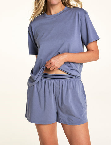 In a softly lit studio, Meagan wears the Cloud Cotton Short and matching Cloud Cotton Tee in Slate. She lifts the hem of the relaxed-fit tee, revealing the logo waistband and button-front detail of the soft cotton modal lounge shorts. sizing: Meagan wears size Small