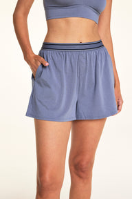 In a softly lit studio, Meagan wears the Cloud Cotton Short in Slate with a matching bralette. The relaxed lounge short features soft cotton modal fabric, side pockets, a button-front detail, and a logo waistband designed for easy everyday comfort.