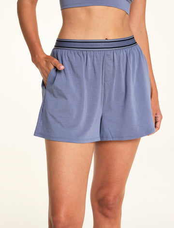 In a softly lit studio, Meagan wears the Cloud Cotton Short in Slate with a matching bralette. The relaxed lounge short features soft cotton modal fabric, side pockets, a button-front detail, and a logo waistband designed for easy everyday comfort.