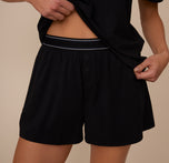 Close-up of Marcela lifting her shirt to reveal the Cloud Cotton Sleep Short in Black. The shorts feature a soft, branded waistband and a mock fly with two buttons for a cozy, lounge-ready look. sizing: Marcela wears size XS