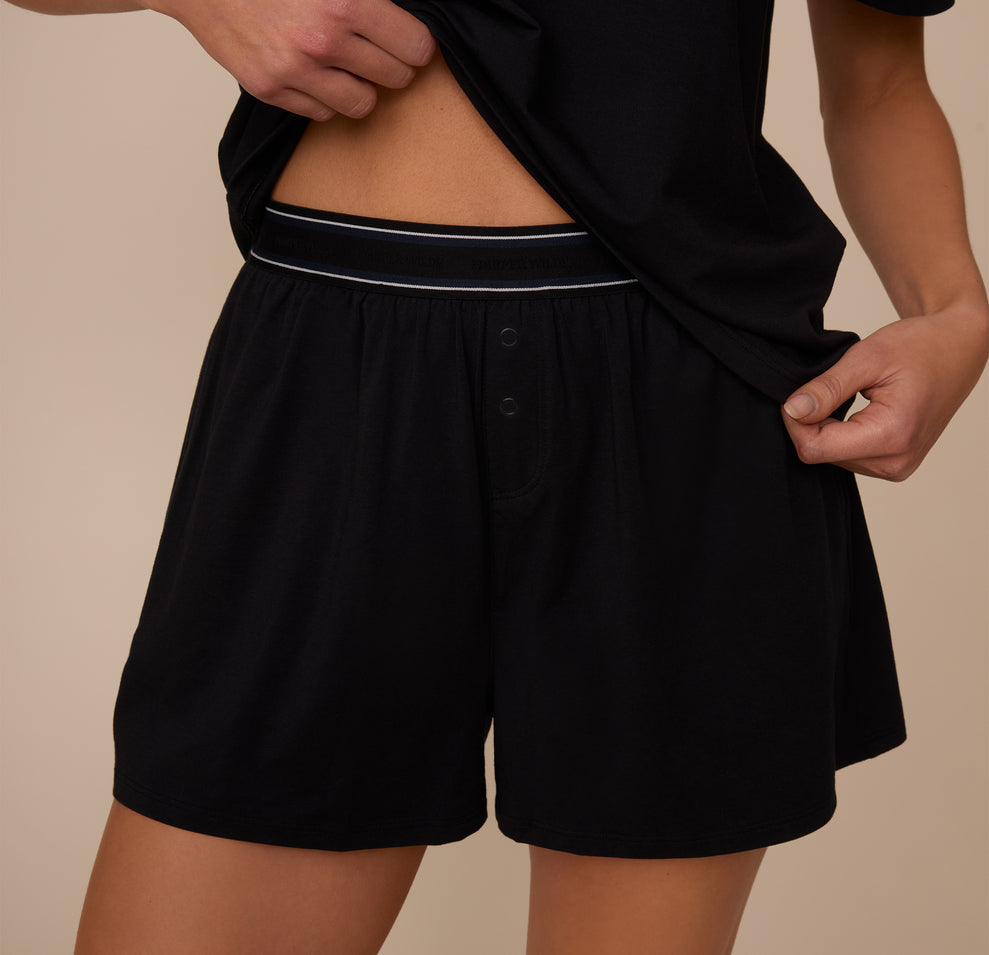 Close-up of Marcela lifting her shirt to reveal the Cloud Cotton Sleep Short in Black. The shorts feature a soft, branded waistband and a mock fly with two buttons for a cozy, lounge-ready look. sizing: Marcela wears size XS
