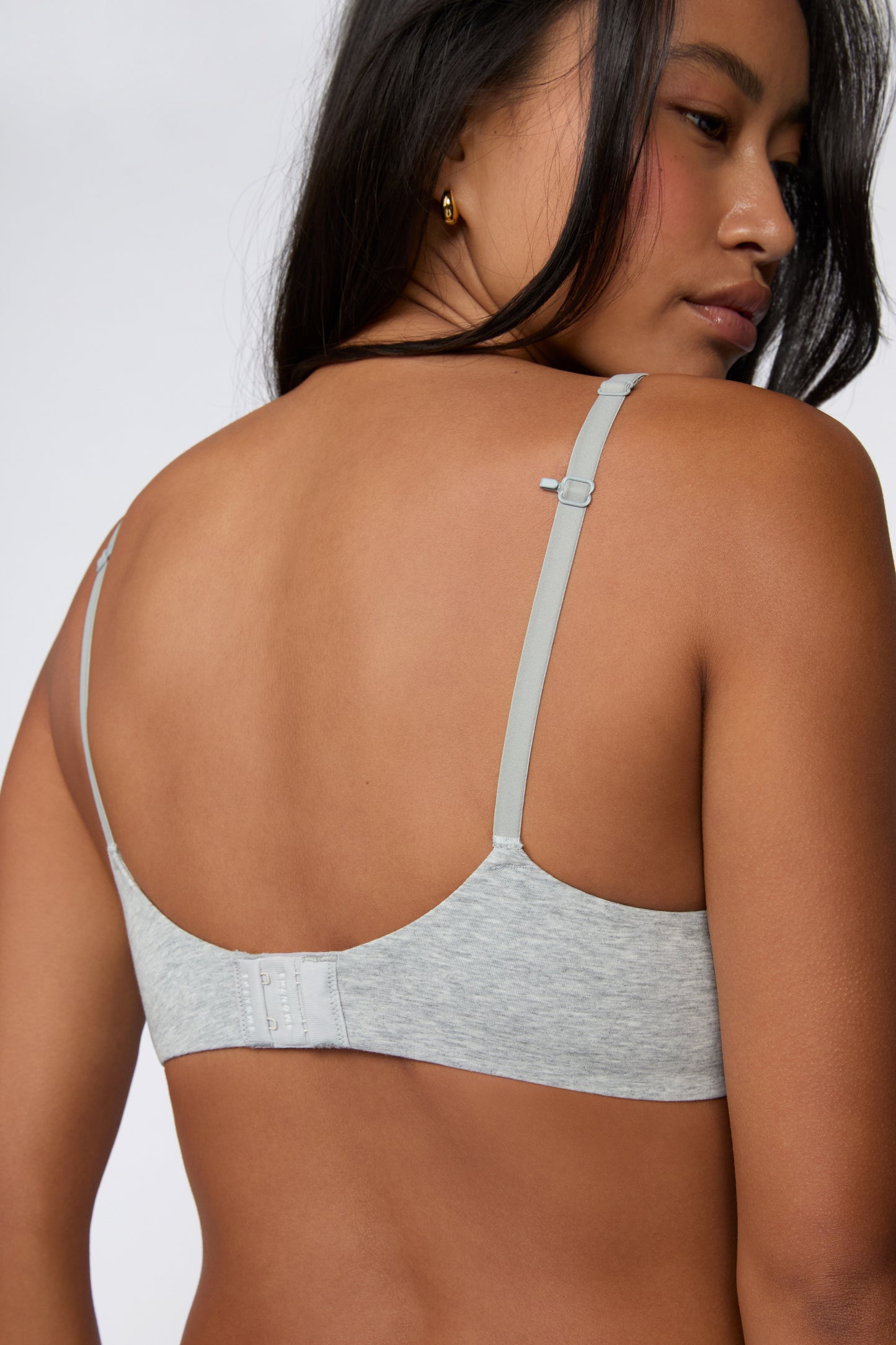 On a white studio backdrop, Amber wears the Cloud Cotton T-Shirt Bra in Heather Grey in a back three-quarter view, showing the adjustable straps, smooth cotton modal band, and low-profile back design.