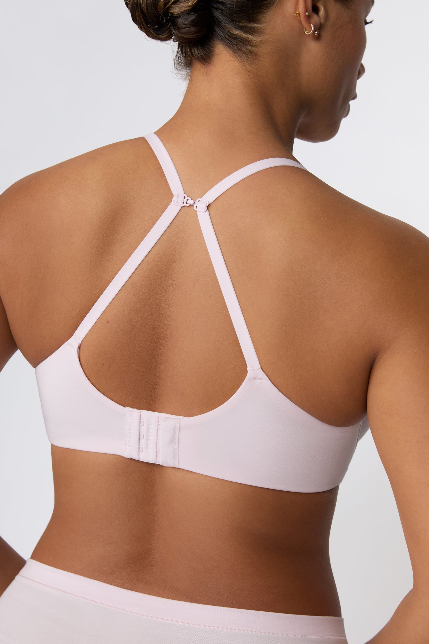 On a white studio backdrop, India is shown from the back wearing the Cloud Cotton T-Shirt Bra in Primrose, highlighting the convertible J-hook straps worn in a racerback position and the hook-and-eye back closure.