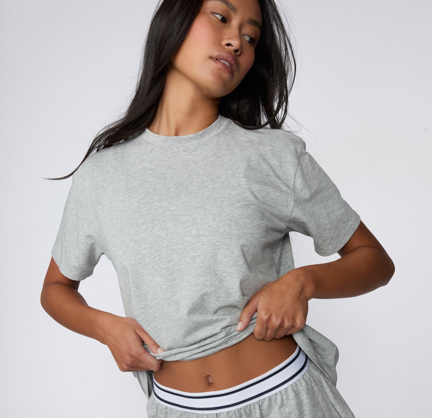On a white studio backdrop, a model wears the Cloud Cotton Tee in Heather Grey, lifting the hem slightly to show the soft cotton fabric and relaxed everyday fit.