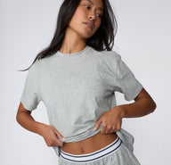 On a white studio backdrop, a model wears the Cloud Cotton Tee in Heather Grey, lifting the hem slightly to show the soft cotton fabric and relaxed everyday fit.