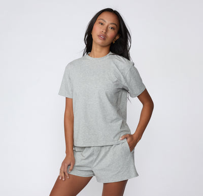 On a white studio backdrop, a model wears the Cloud Cotton Tee in Heather Grey styled with matching lounge shorts, showing the full-length fit and easy drape.