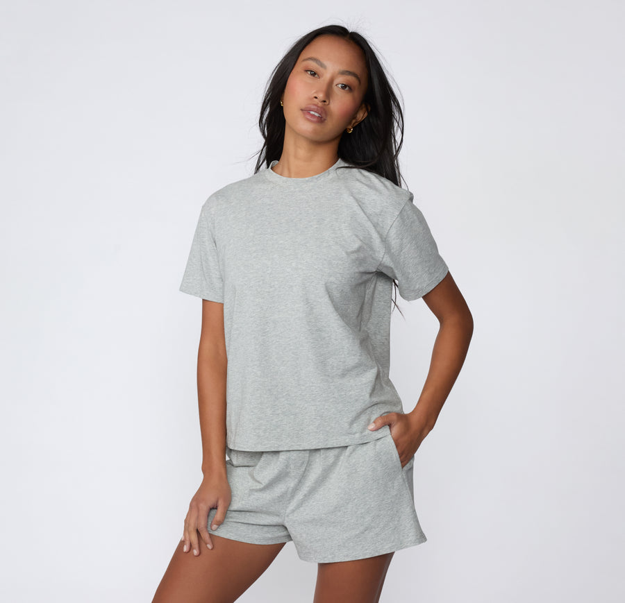 On a white studio backdrop, a model wears the Cloud Cotton Tee in Heather Grey styled with matching lounge shorts, showing the full-length fit and easy drape.