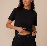 Close-up of Marcela lifting the hem of the Cloud Cotton T-Shirt in Black to reveal the waistband of the matching Cloud Cotton Short. The crewneck tee has a relaxed fit and soft drape, made from breathable cotton-modal fabric. sizing: Marcela wears size XS