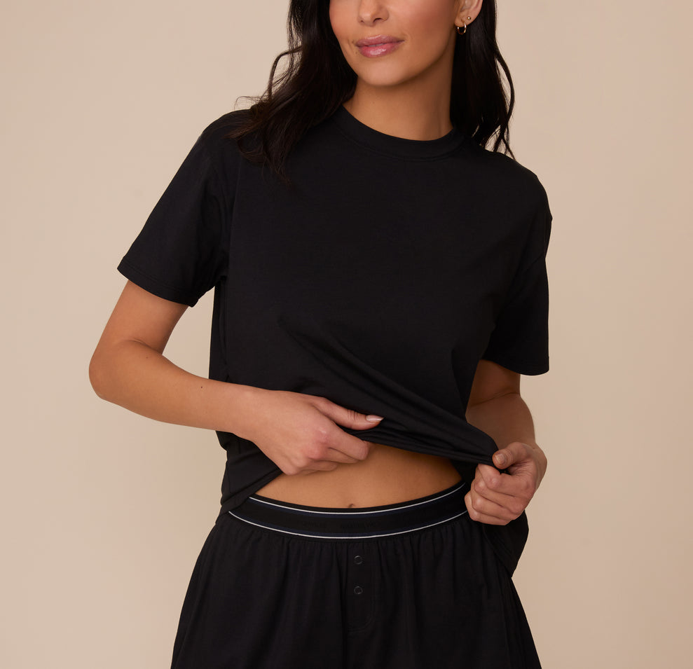 Close-up of Marcela lifting the hem of the Cloud Cotton T-Shirt in Black to reveal the waistband of the matching Cloud Cotton Short. The crewneck tee has a relaxed fit and soft drape, made from breathable cotton-modal fabric. sizing: Marcela wears size XS