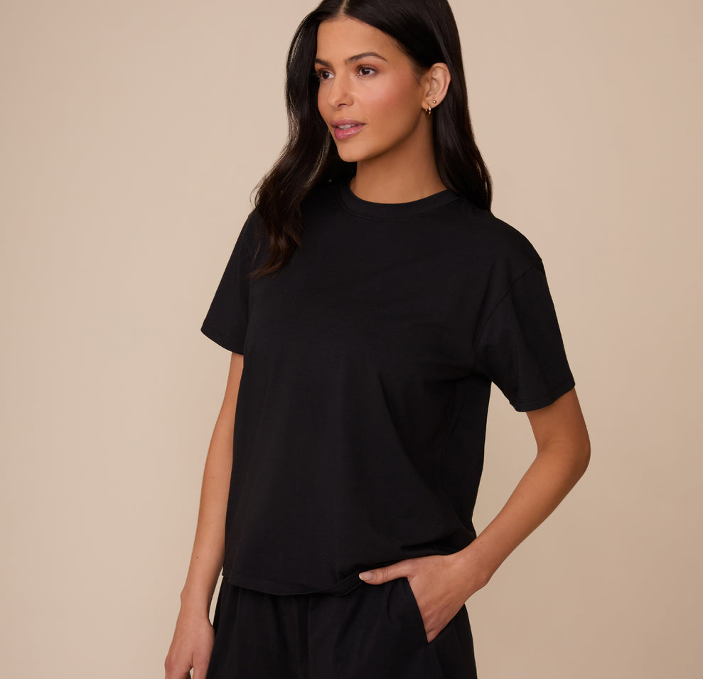 Three-quarter front view of Marcela in the Cloud Cotton T-Shirt in Black. She stands with one hand in her pocket, showing off the relaxed, everyday fit and classic crew neckline of the tee.