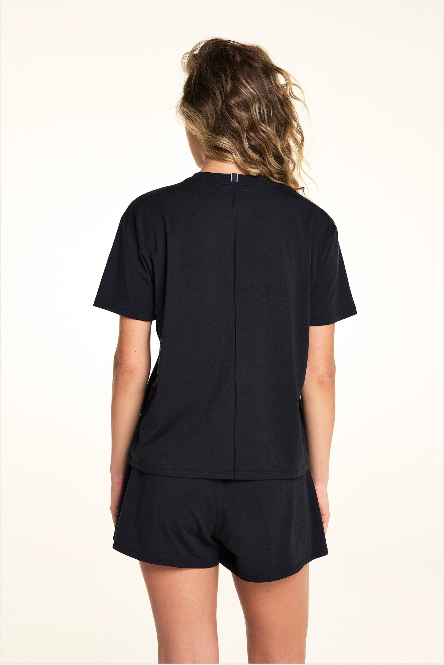 In a studio setting, Alexis wears the Cloud Cotton Tee in Black with matching Cloud Cotton Shorts, shown from the back to highlight the relaxed fit, short sleeves, and soft drape of the fabric.