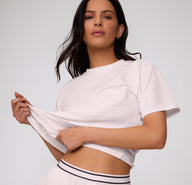 On a clean studio backdrop, a front view shows a model wearing the Cloud Cotton Tee in Primrose, gently stretching the fabric to highlight the soft, flexible cotton modal material.