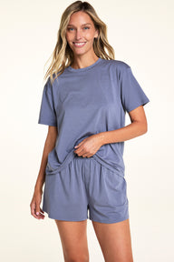 In a softly lit studio, Meagan wears the Cloud Cotton Tee and Cloud Cotton Short in Slate. She lifts the hem of the relaxed-fit tee, revealing the soft cotton modal shorts with side pockets and a button-front detail.