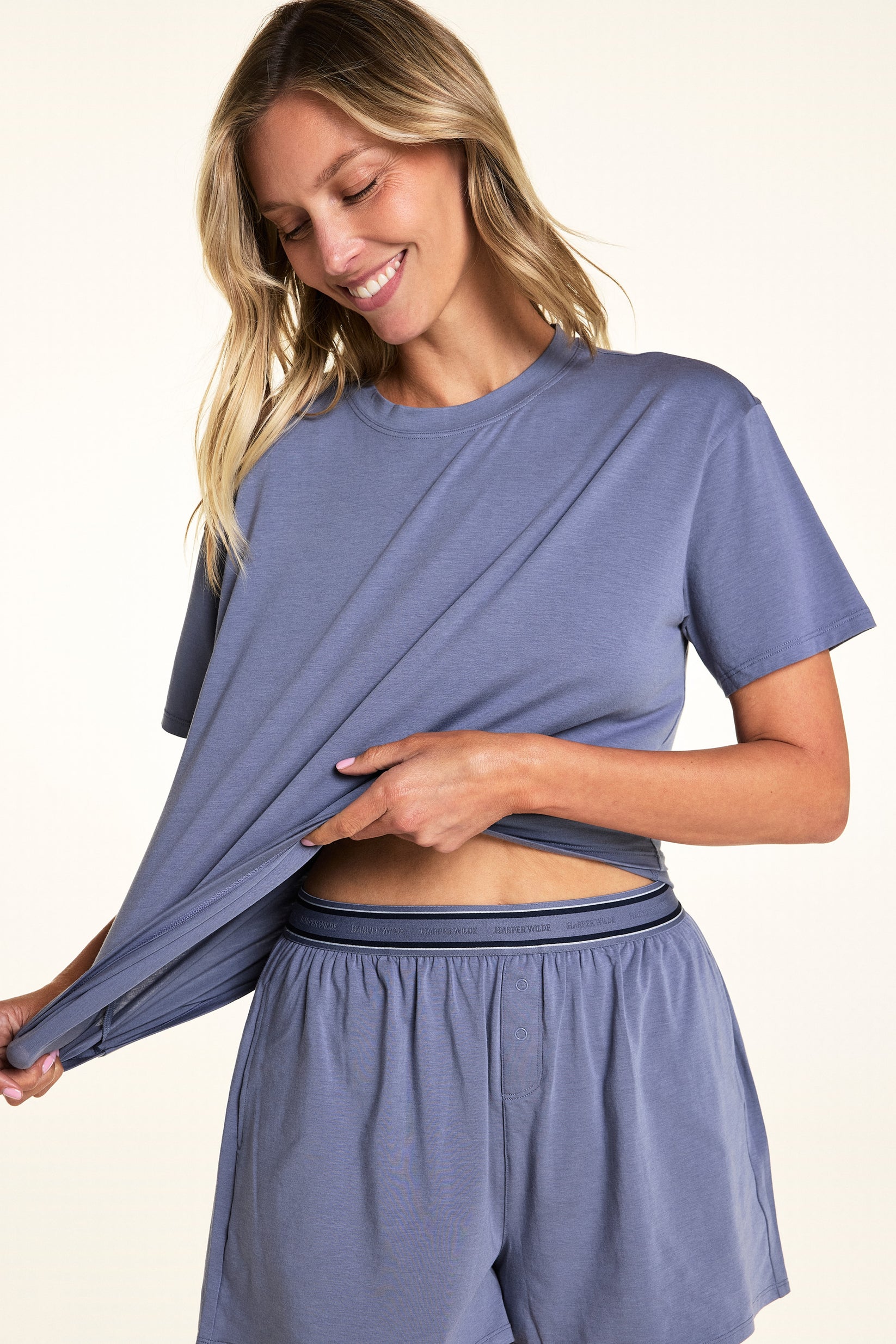 In a softly lit studio, Meagan wears the Cloud Cotton Tee and Cloud Cotton Short in Slate. She stretches the hem of the relaxed-fit tee to show the soft, lightweight cotton modal fabric, paired with lounge shorts featuring a logo waistband and button-front detail. sizing: Meagan wears size Small