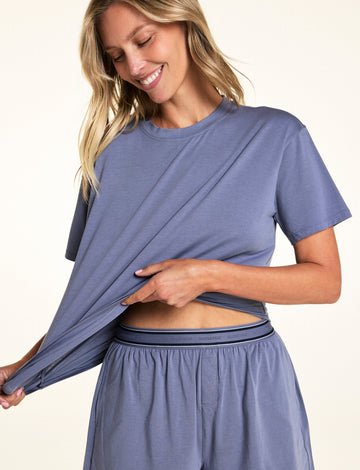 In a softly lit studio, Meagan wears the Cloud Cotton Tee and Cloud Cotton Short in Slate. She stretches the hem of the relaxed-fit tee to show the soft, lightweight cotton modal fabric, paired with lounge shorts featuring a logo waistband and button-front detail. sizing: Meagan wears size Small
