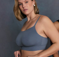 On a textured studio backdrop, a model wears the Cloud Cotton Thin Strap Bralette in Slate, showing the scoop neckline, thin adjustable straps, and smooth cotton-modal fabric. The wireless design and soft underband are visible, highlighting comfortable everyday support.
