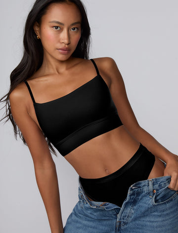 On a grey studio backdrop, Amber wears the Cloud Cotton Thong in Black with relaxed denim and a matching black bralette, featuring soft cotton modal fabric and a smooth, minimal coverage silhouette. sizing: Amber wears size Small