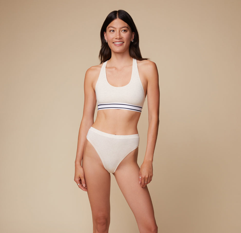 Anna stands facing the camera, wearing the Cloud Cotton Highwaist Thong in Heather Oatmeal with a matching Cloud Cotton Logo Racerback Bralette. The high waistband of the thong is emphasized. sizing: Anna wears size Small