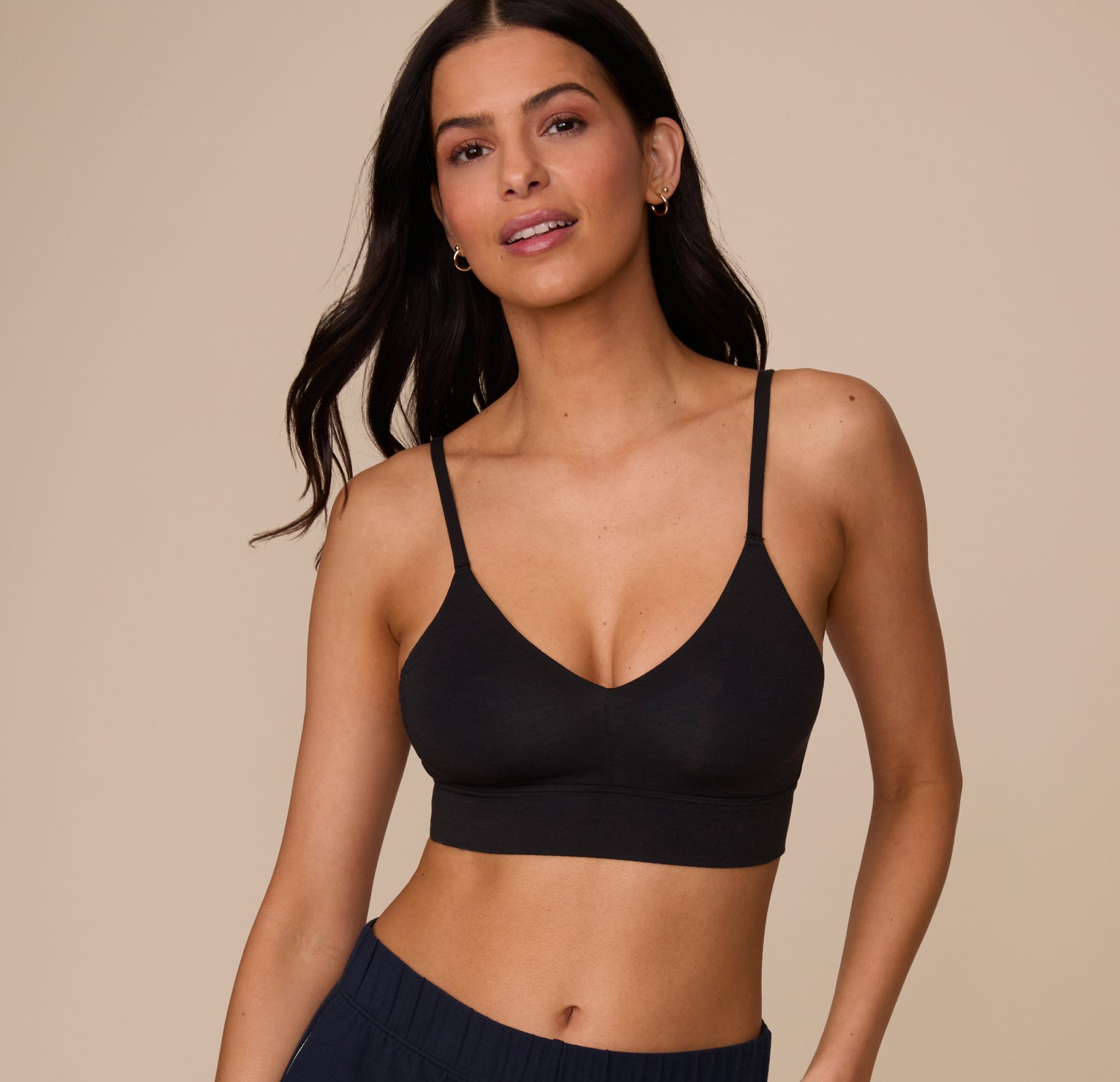 Against a warm neutral backdrop, a model wears the Cloud Cotton Triangle Bralette in Black, shown from the waist up. The bralette features a soft triangle shape with a wide underband and adjustable straps, made from breathable cotton fabric for comfortable, everyday wear.