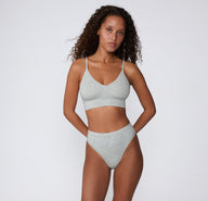 On a light studio background, a model stands wearing the Cloud Cotton Triangle Bralette in Heather Grey with the matching Cloud Cotton Bikini, showing the wireless triangle shape, thin adjustable straps, and comfortable longline fit.