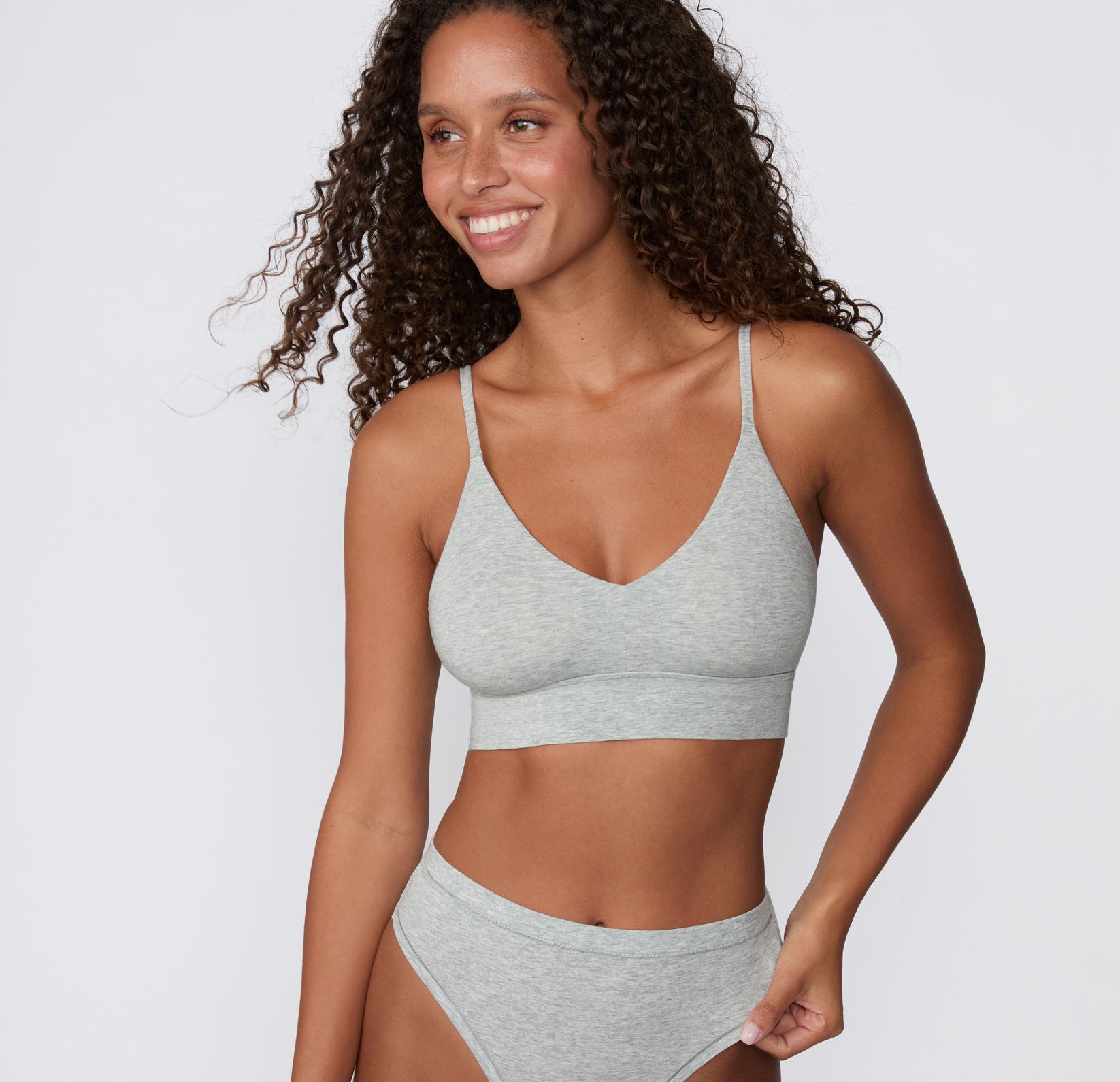 On a light studio backdrop, a model wears the Cloud Cotton Triangle Bralette in Heather Grey with the matching Cloud Cotton Bikini, showing the wireless triangle shape, thin adjustable straps, and soft longline underband.