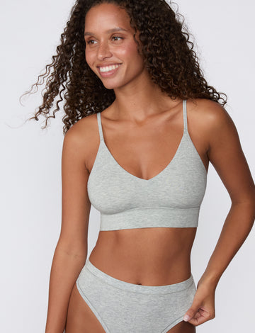 On a light studio backdrop, a model wears the Cloud Cotton Triangle Bralette in Heather Grey with the matching Cloud Cotton Bikini, showing the wireless triangle shape, thin adjustable straps, and soft longline underband.