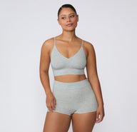 On a light studio background, a model wears the Cloud Cotton Triangle Bralette in Heather Grey with matching Cloud Cotton shorts, showing the wireless triangle shape, longline underband, and lightweight cotton-modal construction.
