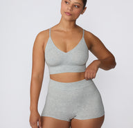 On a clean studio backdrop, a model wears the Cloud Cotton Triangle Bralette in Heather Grey with matching Cloud Cotton shorts, showing the wireless triangle silhouette, thin adjustable straps, and soft longline underband.