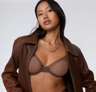 On a white studio backdrop, a model wears the ComfortMesh™ Plunge Bra in Cashmere Amber, styled under an open brown leather jacket, showing the sheer mesh cups and plunging neckline.