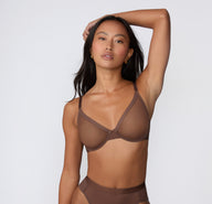 On a white studio backdrop, a model wears the ComfortMesh™ Plunge Bra in Cashmere Amber, raising one arm to show the plunging front, sheer mesh fabric, and adjustable straps.