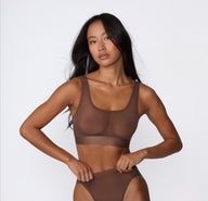 On a white studio backdrop, Amber wears the ComfortMesh™ Scoop Bralette in Cashmere with matching bottoms, shown in a front-facing three-quarter view highlighting the sheer mesh fabric, scoop neckline, and wide supportive band.