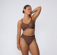 On a white studio backdrop, India wears the ComfortMesh™ Triangle Bralette in Cashmere with matching bottoms, shown in a full-body view highlighting the sheer mesh fabric, V-neckline, adjustable straps, and wide supportive band.
