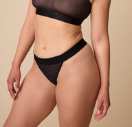 Emersyn poses wearing the ComfortMesh Thong in Black. The wide, comfortable waistband and sheer, stretchy fabric are emphasized. sizing: Emersyn wears a size Medium