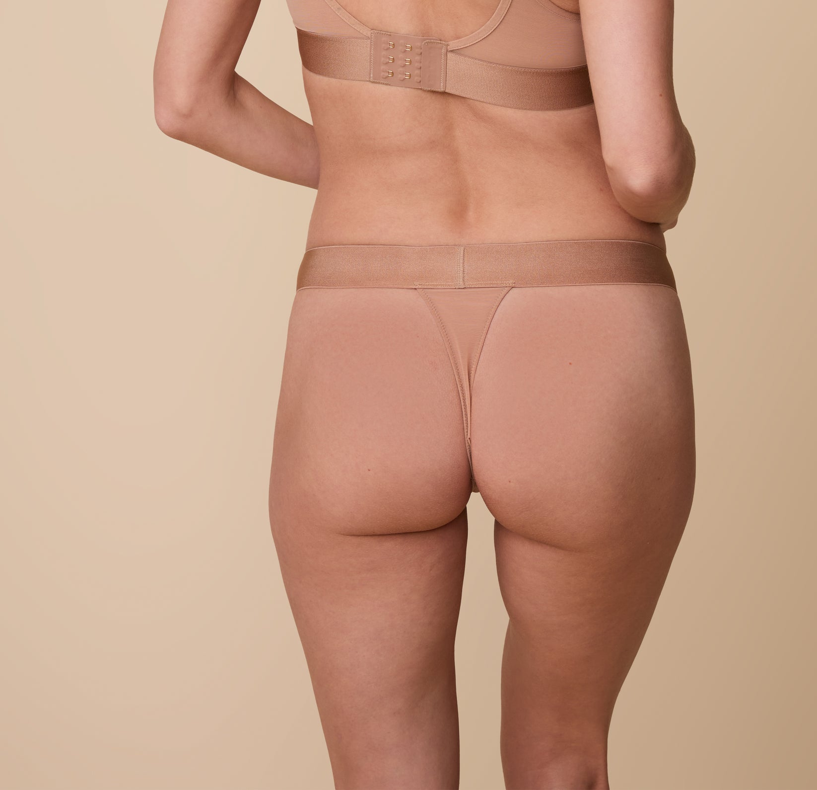 Gracie from the back, wearing the ComfortMesh™ Thong in Taupe, displaying the thong’s minimal back coverage and seamless wide waistband that lays flat against the skin. The image highlights the thong’s comfortable fit.