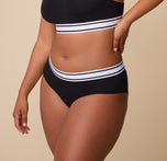 Three quarters view of the Cloud Cotton Retro Hiphugger in Black. The model places her hand on her hip, highlighting the high-rise fit and curve-hugging design.