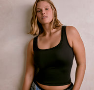 Against a neutral indoor wall, a model wears the Flex Racerback Sport Tank in Black. The fitted tank features a smooth, supportive fabric with a scoop neckline and racerback design, styled casually with denim for an everyday, wear-anywhere look.