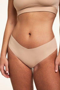 Close-up on a light studio backdrop of Sonnie wearing the Invisible No-Show Low Rise Thong in Beige with a matching scoop bralette. The thong features a low-rise silhouette with elastic-free edges for a smooth, second-skin feel.