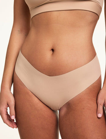 Close-up on a light studio backdrop of Sonnie wearing the Invisible No-Show Low Rise Thong in Beige with a matching scoop bralette. The thong features a low-rise silhouette with elastic-free edges for a smooth, second-skin feel.