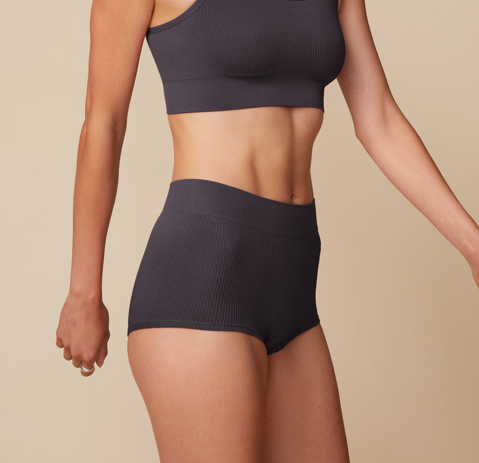 Side view of Anna wearing the Lounge Rib Scoop Bralette and Boyshort in Charcoal. The matching set emphasizes the comfort and every day wearability of the products.