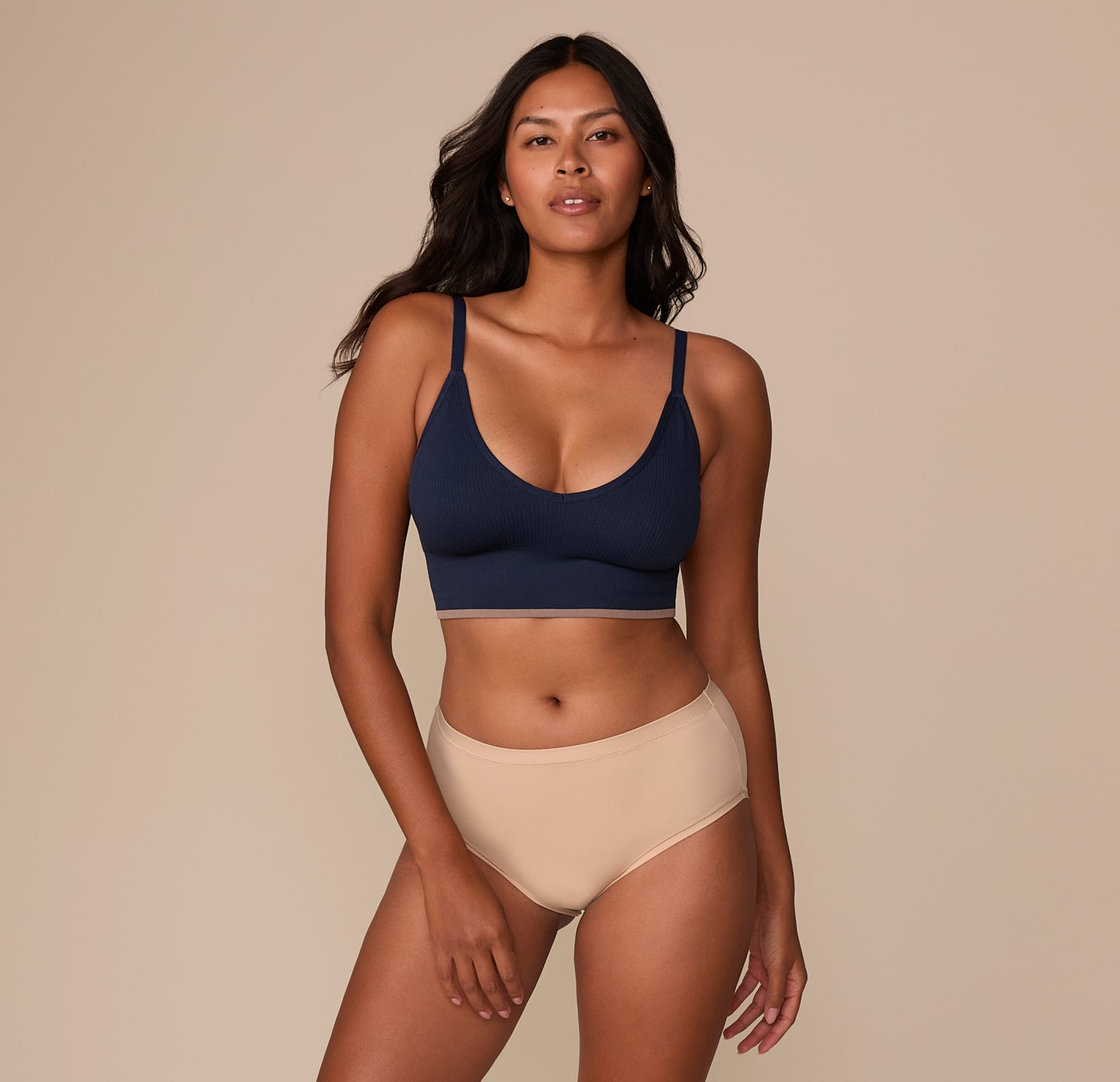 Victoria stands confidently in a full-body shot wearing the Lounge Rib Triangle Bralette in True Blue, paired with the Bliss Highwaist Brief in Beige. The bralette's sleek silhouette and modern contrast hem highlight comfort with style.