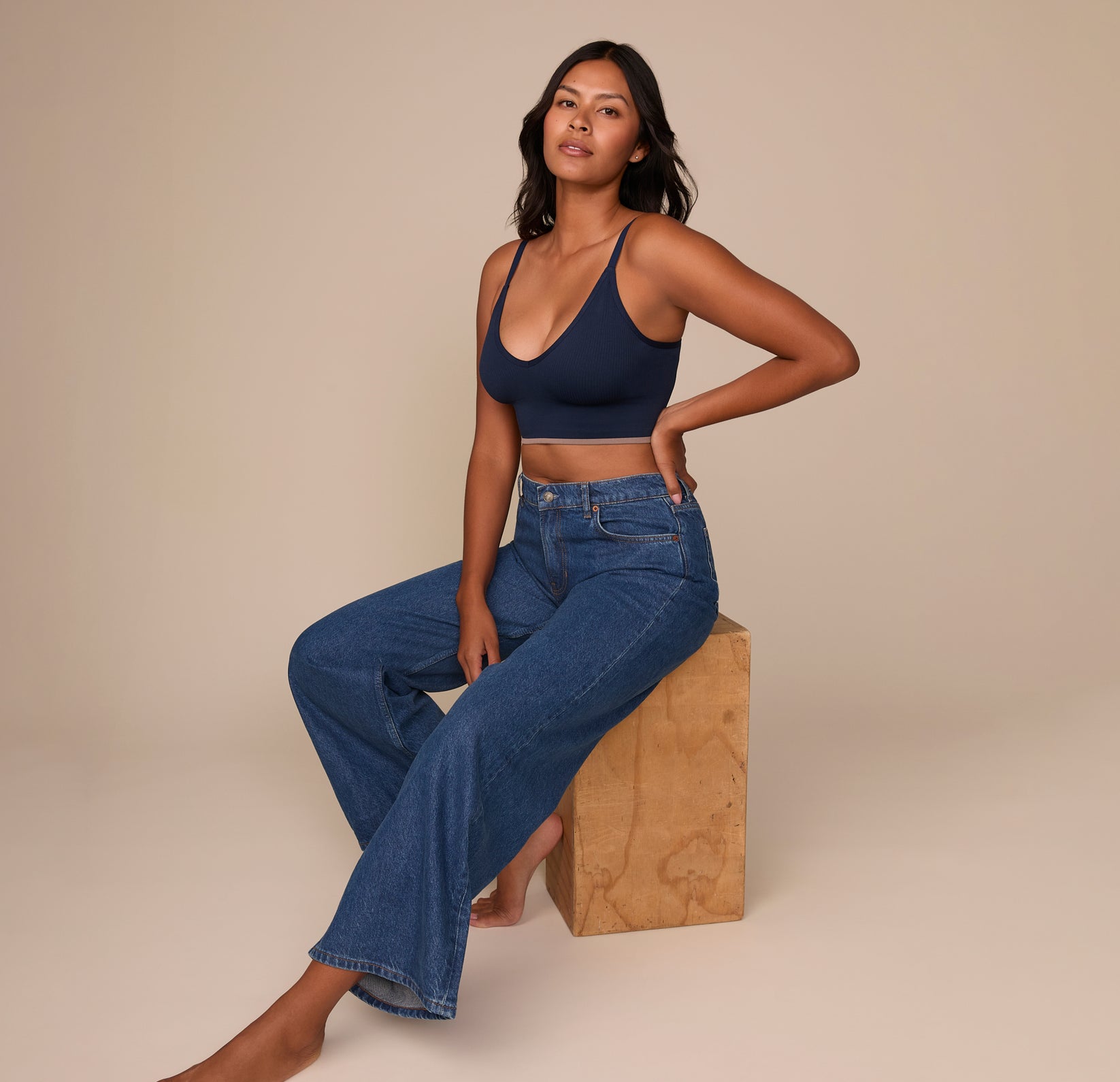Victoria sits on a wooden block, styled in wide-leg jeans and the Lounge Rib Triangle Bralette in True Blue. This lifestyle image captures the bralette’s versatile wearability beyond loungewear, styled casually for everyday comfort.