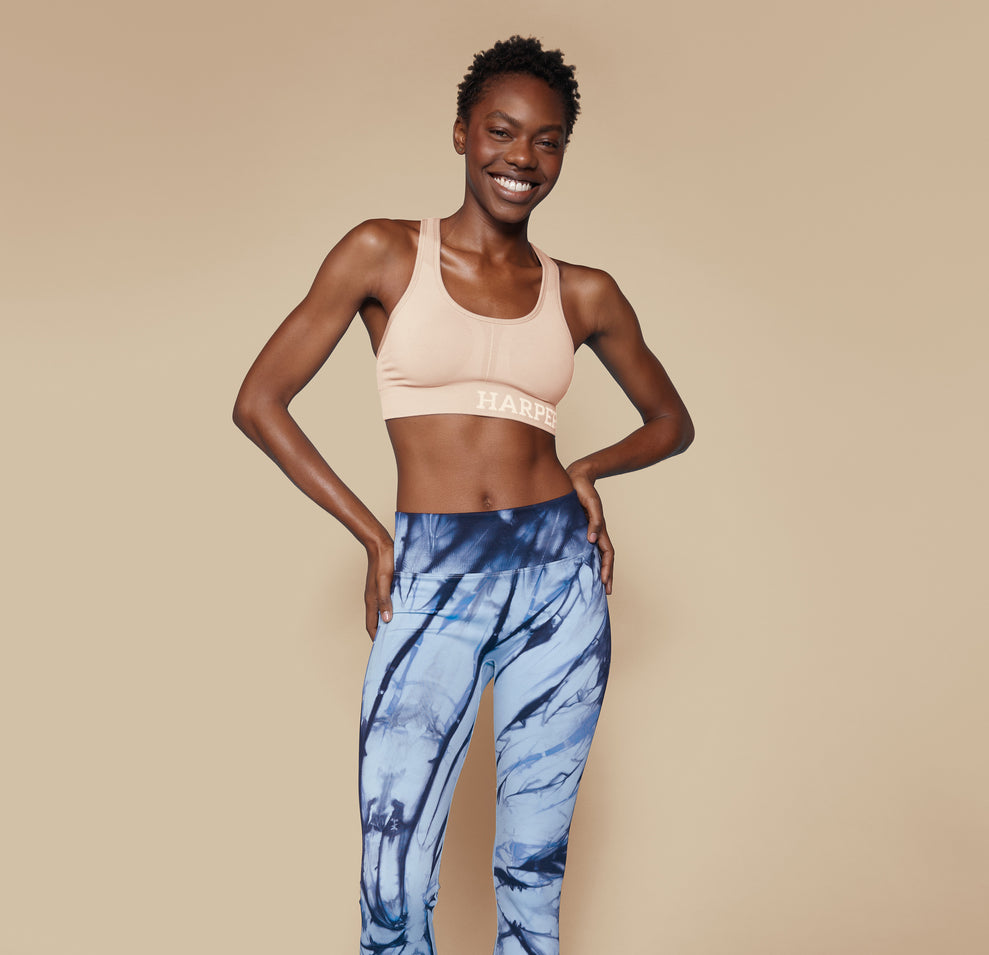 Full body shot of Mariah in the Move Sports Bra in Beige, paired with blue tie-dye leggings, standing with hands on hips and smiling confidently.