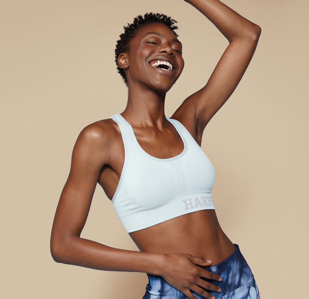 Model Mariah smiles brightly while wearing the Move Sports Bra in Ice, with one arm raised, showcasing the sporty fit and Harper Wilde logo on the band.