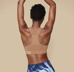 Back view of model Mariah in the Move Sports Bra in Tan, with both arms raised, displaying the racerback style.