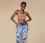 Full body shot of Mariah in the Move Sports Bra in Tan, paired with blue tie-dye leggings, standing with hands on hips and smiling confidently.