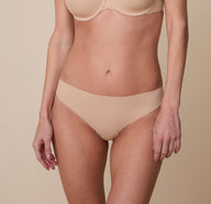 Gracie wearing the No-Show Thong in Beige. The smooth, seamless waistband sits flat against her skin, providing a comfortable and invisible look under clothing. sizing: Gracie wears size XS