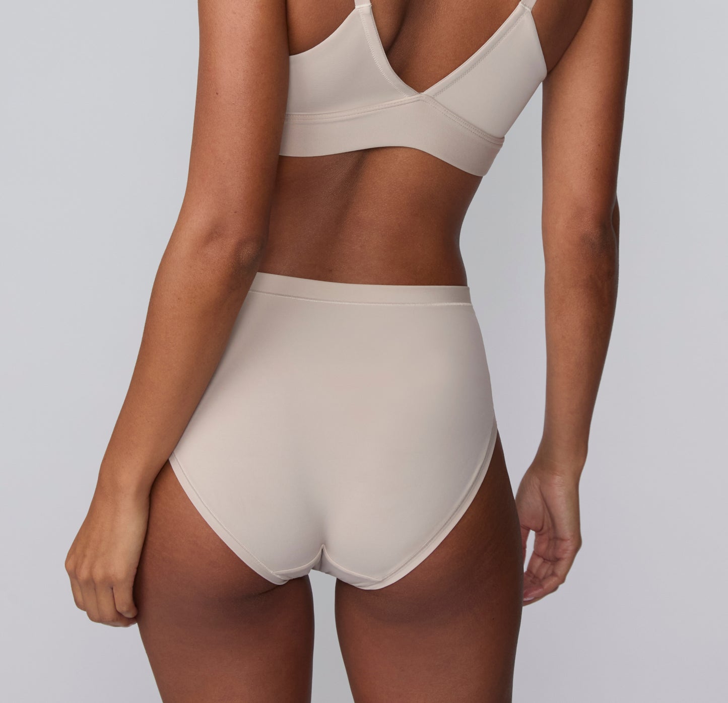 On a light gray studio backdrop, Courtney wears the Bliss Highwaist Brief in Pebble with the matching Bliss Triangle Bralette, shown from the back highlighting the high-rise waist, smooth microfiber fabric, and clean back coverage.