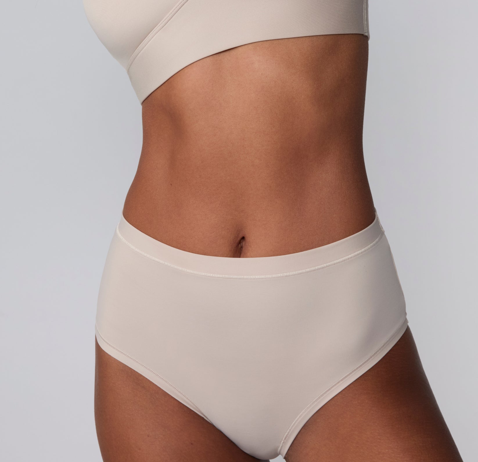 On a light gray studio backdrop, Courtney wears the Bliss Highwaist Brief in Pebble, shown in a close-up front crop highlighting the smooth microfiber fabric, high-rise waistband, and clean, no-show edges at the waist.
